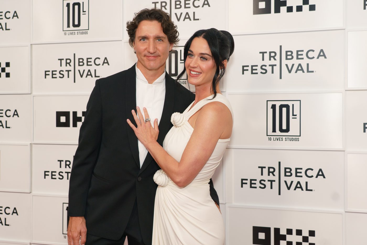 A man in a black suit and a woman in a white dress pose together, smiling, in front of a backdrop with “Tribeca Festival” and various logos.